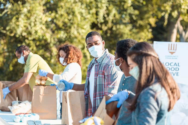 A mid adult man places a canned food item into a paper bag as he volunteers with his teenage son at an outdoor food bank. He and other volunteers are wearing protective face masks as they are volunteering during the coronavirus pandemic.